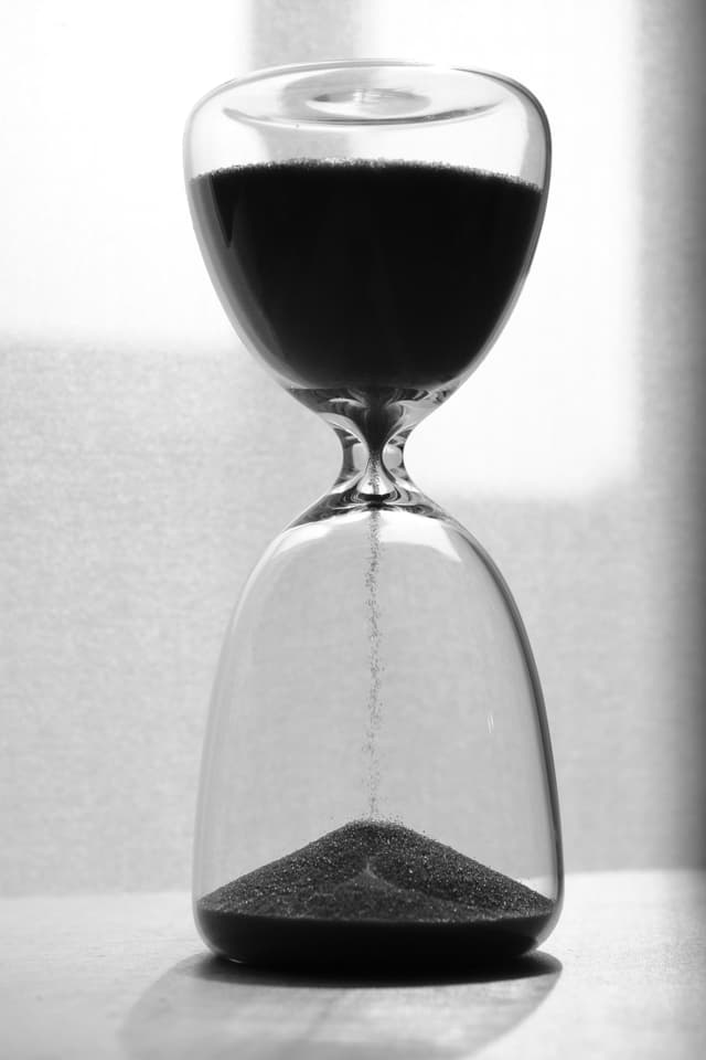 A black and white studio photograph of a glass hourglass with dark sand trickling from the top bulb into the base