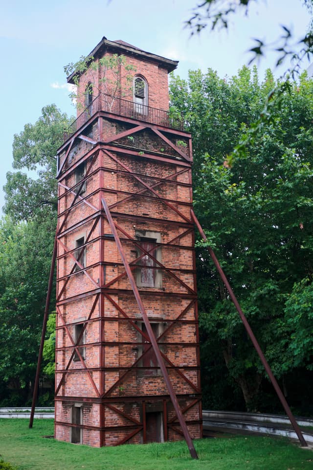 Tall brick tower with external metal framework, surrounded by green trees and grass