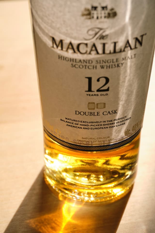 Close-up of a Macallan 12 Year Old Double Cask Scotch whisky bottle showing the label and glowing golden liquid against a light surface
